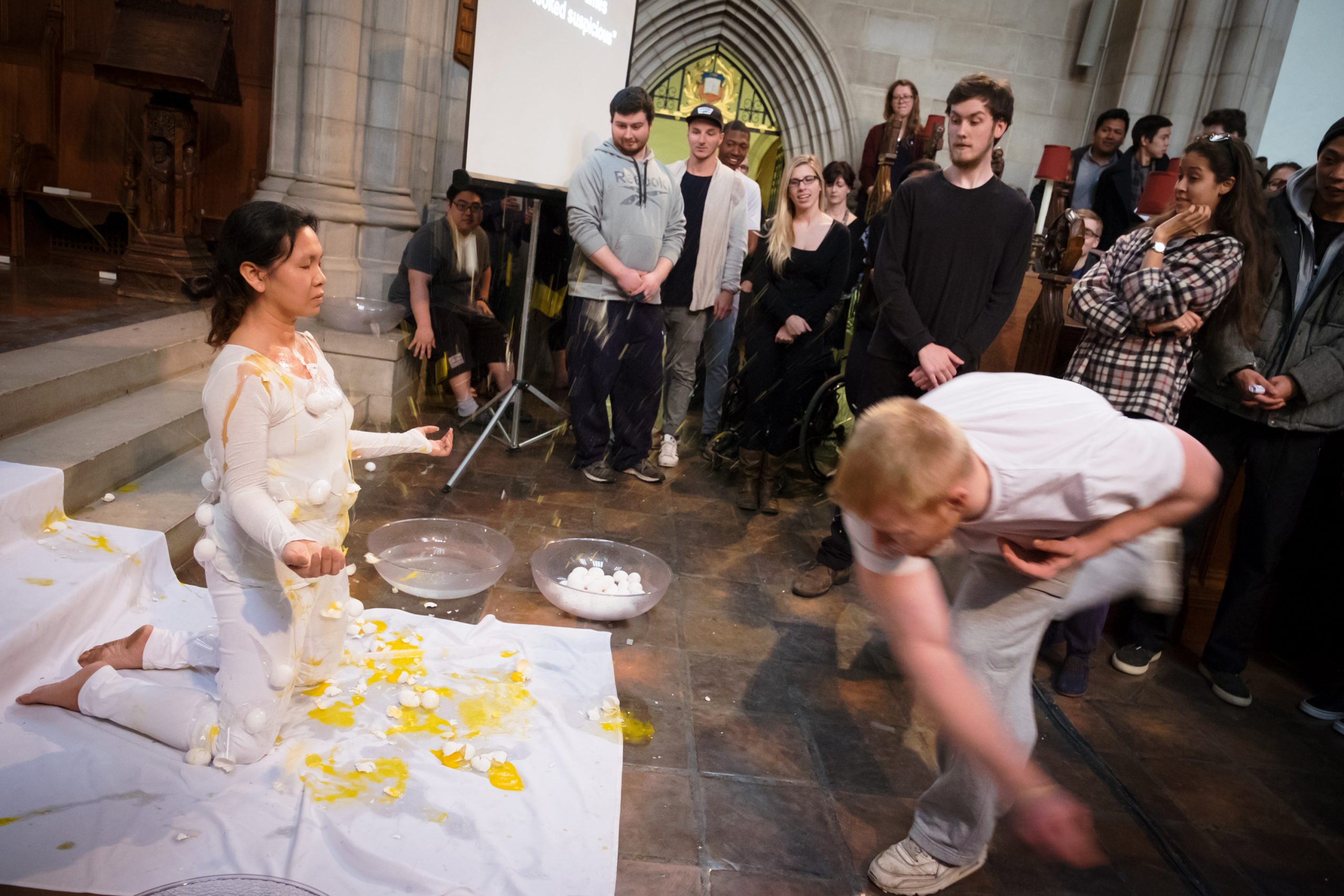 In this action shot inside of Trinty College Chapel, a white male student throws an egg at artist Anida Yoeu Ali as a group of students look on. Anida wears a white bodysuit and is kneeling on a white cloth runner. Her eyes are closed and her arms at her sides with her palms facing up in a meditative surrender. She is covered with broken eggshells and splattered yolks. Two large clear plastic bowls are by her left side - one is filled with water and the other with remaining eggs.