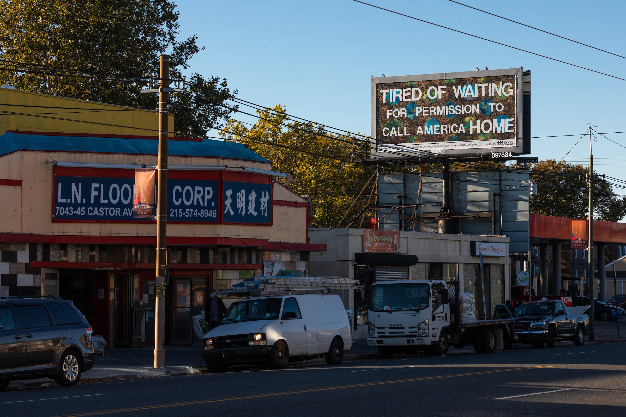A sunlit billboard in Philadelphia is the main focus of this horizontally oriented photograph. Written in large white block letters against a busy abstract patterned background, the sign reads “Tired of Waiting for Permission to Call America Home.” According to Mural Arts Philidelphia’s Instagram, this was billboard is part of their “Messages to Our Neighbors” project in which artist Aram Han Sifuentes “worked with high school students to explore the intersection of citizenship, immigration, and belonging.” The billboard hangs between powerlines and there are trees in the background of this Northeast Philadelphia street scene. The Castor Avenue street below is a quiet multicultural urban street with a flooring business with Chinese lettering, a hair salon, and a coffee shop. Work trucks and vans are parked in front.