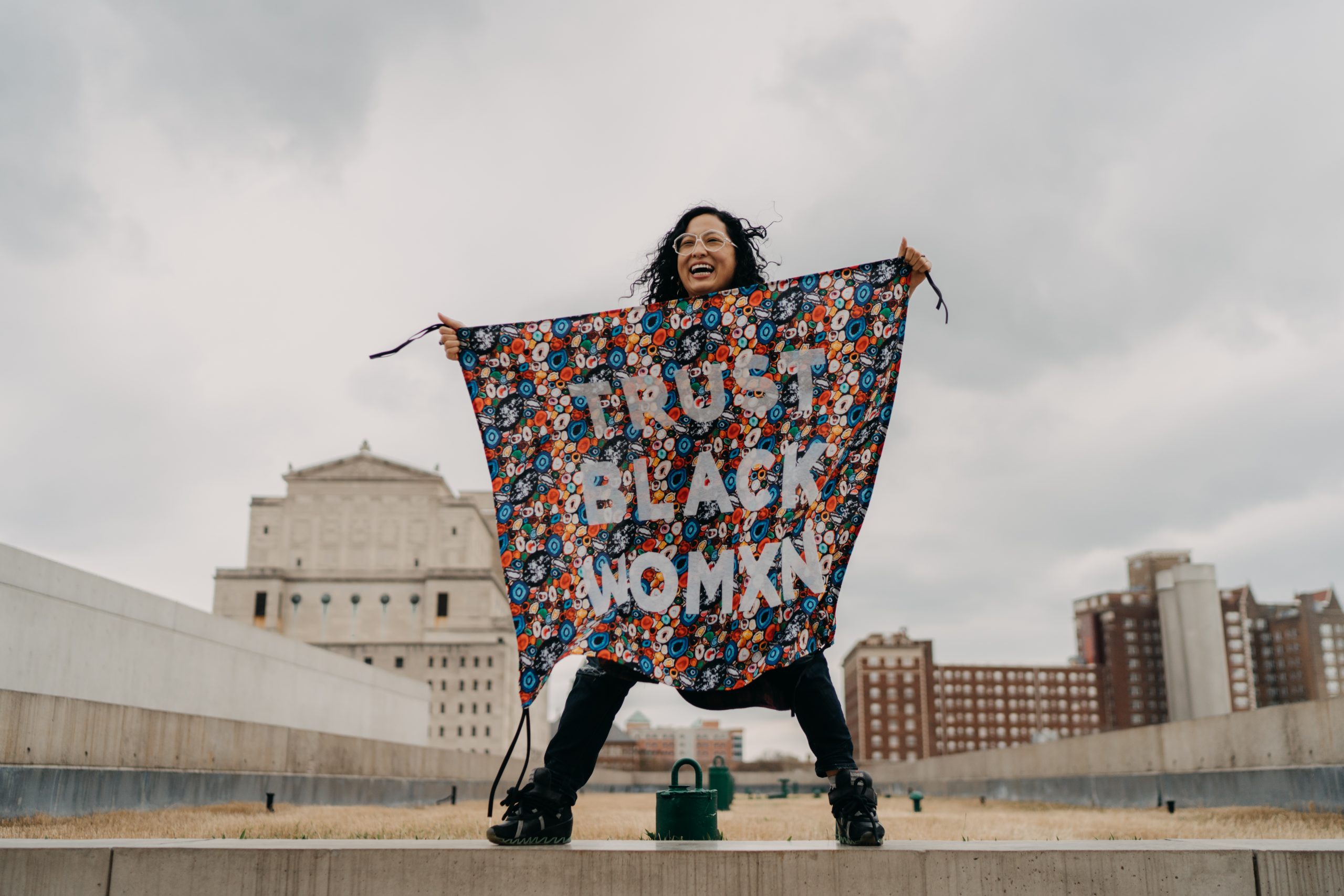Aram Han Sifuentes is pictured standing on the rooftop of the Pulitzer Arts Foundation in St. Louis, Missouri. With an exuberant smile on her face, she has her feet spread far apart and her arms are outstretched holding a banner with bold white block letters against a busy abstract pattern that reads “Trust Black Womxn.” The banner is from her Protest Banner Lending Library and covers most of her body. Aram, a Korean American woman in her mid-thirties, has wavy long black hair and is dressed in black and wears white polygon-shaped wire-rim glasses.