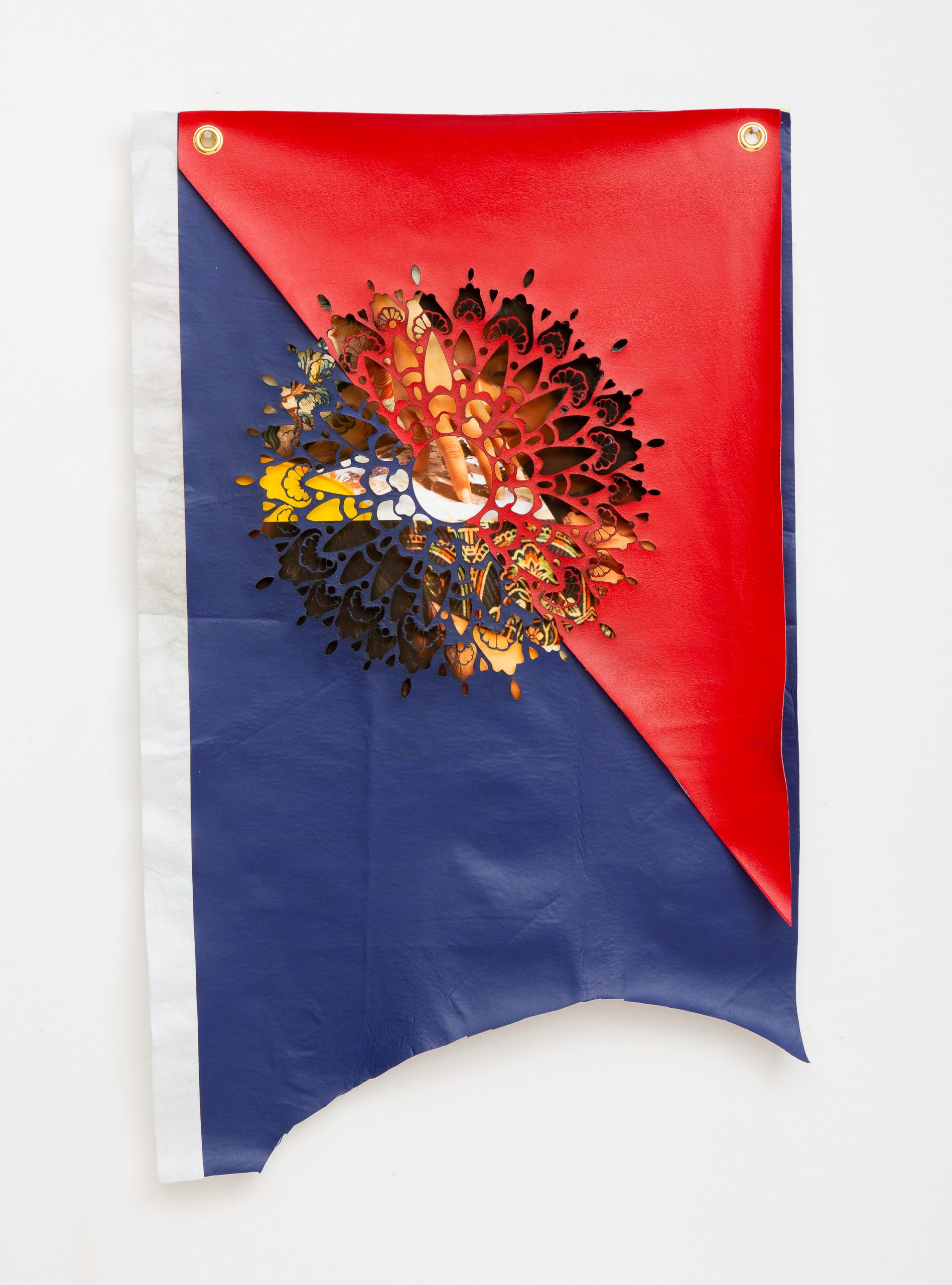 A vertically oriented mixed-media banner/flag hangs from two gold grommets. The top section has a diagonal triangle of red pleather and the bottom section has a diagonal triangle of navy blue pleather. A strip of white runs down the left side. A rounded shape has been roughly cut from the bottom of the banner. In the center of the banner is an ornate circular cutout, which can be seen as a seal or portal. Behind the cutout is a photographic collage of fragments of patterned fabric that also reads as tattooed flesh, the artist’s face, and their hand in the act of arranging materials into a composition.