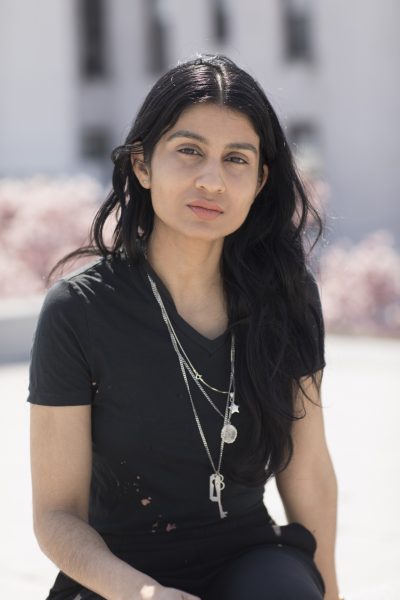 Headshot of Baseera Khan. In this three-quarter view portrait, a self-identified “queer femme Muslim” with peach-colored lipstick and long black wavy hair parted in the middle is standing in the sun looking calmly at the camera with their head slightly tilted. They wear black pants and a black v-neck t-shirt and small silver earrings and three long silver chains with a star, lock and key, and a circular pendant.