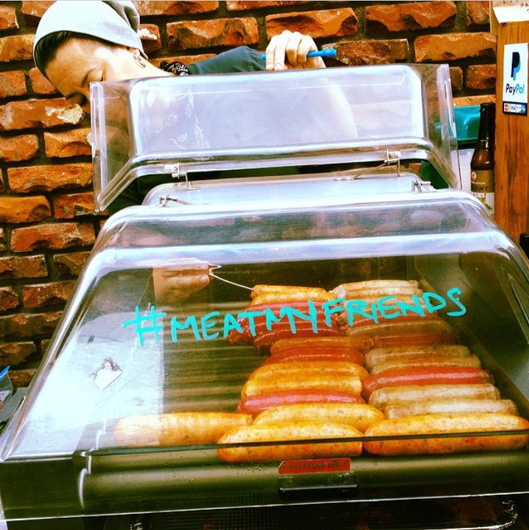 O’Brien lifts the lid of a grillah deck to check the temperature of a sausage. A variety of sausages are featured in two rows on the grill. “#MEATMYFRIENDS” is hand written in turquoise marker on the front of the grill’s clear plastic cover.