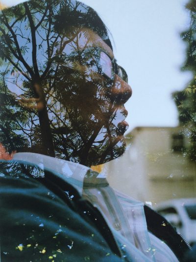 Headshot of Francis Wong. A young Chinese American man with short hair is pictured in profile wearing black-rimmed glasses. He wears a striped white collared shirt and a light-weight blue zippered jacket. The background is a blurred fragment of a building and car. His image is superimposed and blurred artistically with a photo of a treetop canopy.