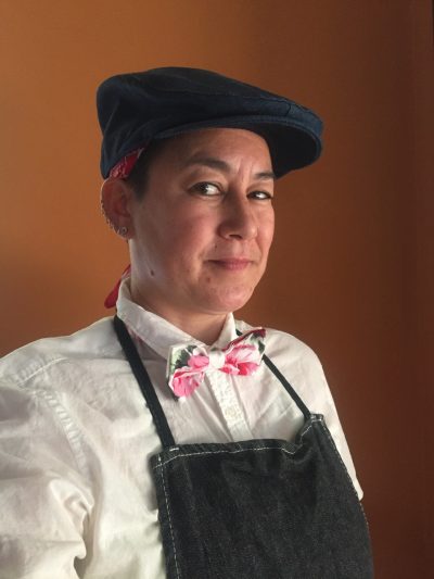 Headshot of Genevieve Erin O’Brien. A middle-aged non-binary mixed-race Vietnamese American person looks at the camera with one eyebrow raised and a slight smile in this three quarter view portrait. They wear a white button-down shirt, bowtie with a pink floral print, a black denim apron, and a matching driver’s cap. Under the cap, a small bit of a red bandana is visible. They have multiple ear piercings and stand against a dark orange wall with directional sunlight.
