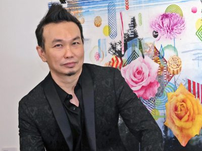 Heinrich Toh headshot. An Asian American man in his forties is seated in front of one of his large-scale prints with colorful and decorative flowers and geometric shapes in the background. He leans in, looking directly at the camera with a slight smile and a twinkle in his eye. He has a goatee and his black hair is styled in a faux hawk. He wears a black floral jacquard dress suit jacket with a black collared shirt with the top two buttons unbuttoned.