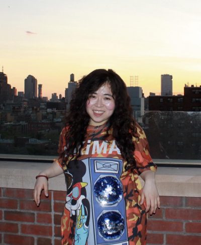 Headshot of HyeGyeong. In this candid portrait, a Korean woman in her mid-thirties with a big smile and long wavy brown hair leans her arms back against a brick rooftop patio ledge. The sun is setting on this cityscape. She wears an oversized earth-toned camouflage t-shirt with a cartoon image of Mickey Mouse with sunglasses chilling out next to a glitter-filled loud speaker.