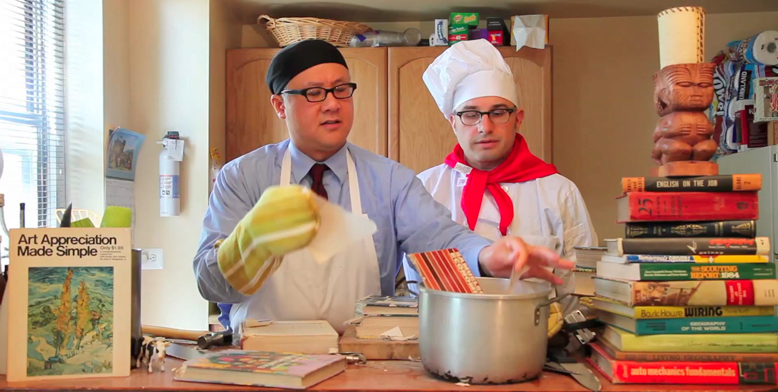 A video still of artists Larry Lee and Jason Dunda in Jason’s kitchen literally cooking books in a mock cooking show skit whose characters are based vaguely on the 1980’s PBS cooking star Martin Yan from Yan Can Cook and the Muppet Show’s Swedish Chef. Larry is a Chinese American man dressed in a blue button-down shirt with a maroon tie and a white apron and he wears a black skull cap and black glasses. Larry wears a yellow mitt and is adding torn pages into a silver soup stockpot in which an economic book is cooking on the counter in front of them. In the foreground to the left of the screen image is an Art Appreciation Made Simple book and a small figurine of a milk cow. Jason, who is a white Canadian man, looks on wearing black glasses, a white chef’s apron and hat, and a red scarf. A tall stack of old instruction manuals with titles such as English on the Job and Auto Mechanics Fundamentals is piled on the counter to the screen right of the duo. On top of the stack of books is a Tiki-style lamp. 