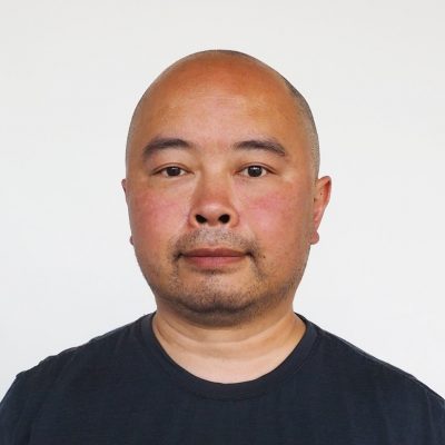 Headshot of Jave Yoshimoto. A middle-aged Chinese American man from Japan with a shaved head and 5’O’clock shadow stubble wears a black t-shirt against a white background. He looks directly at the camera with a slight smile.