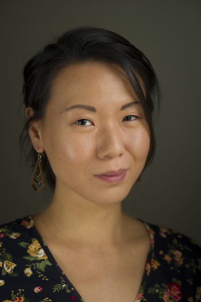Kathy Liao headshot. A Taiwanese American woman in her mid-thirties has a slight smile and twinkle in her eye in this three-quarter view headshot. She has short black bob haircut that is tied back. Loose strands of hair frame her face. She wears bronze drop earrings with a chevron pattern and wears a black v-neck blouse with a yellow and pink floral pattern. The background of this studio photo is a muted dark olive green.