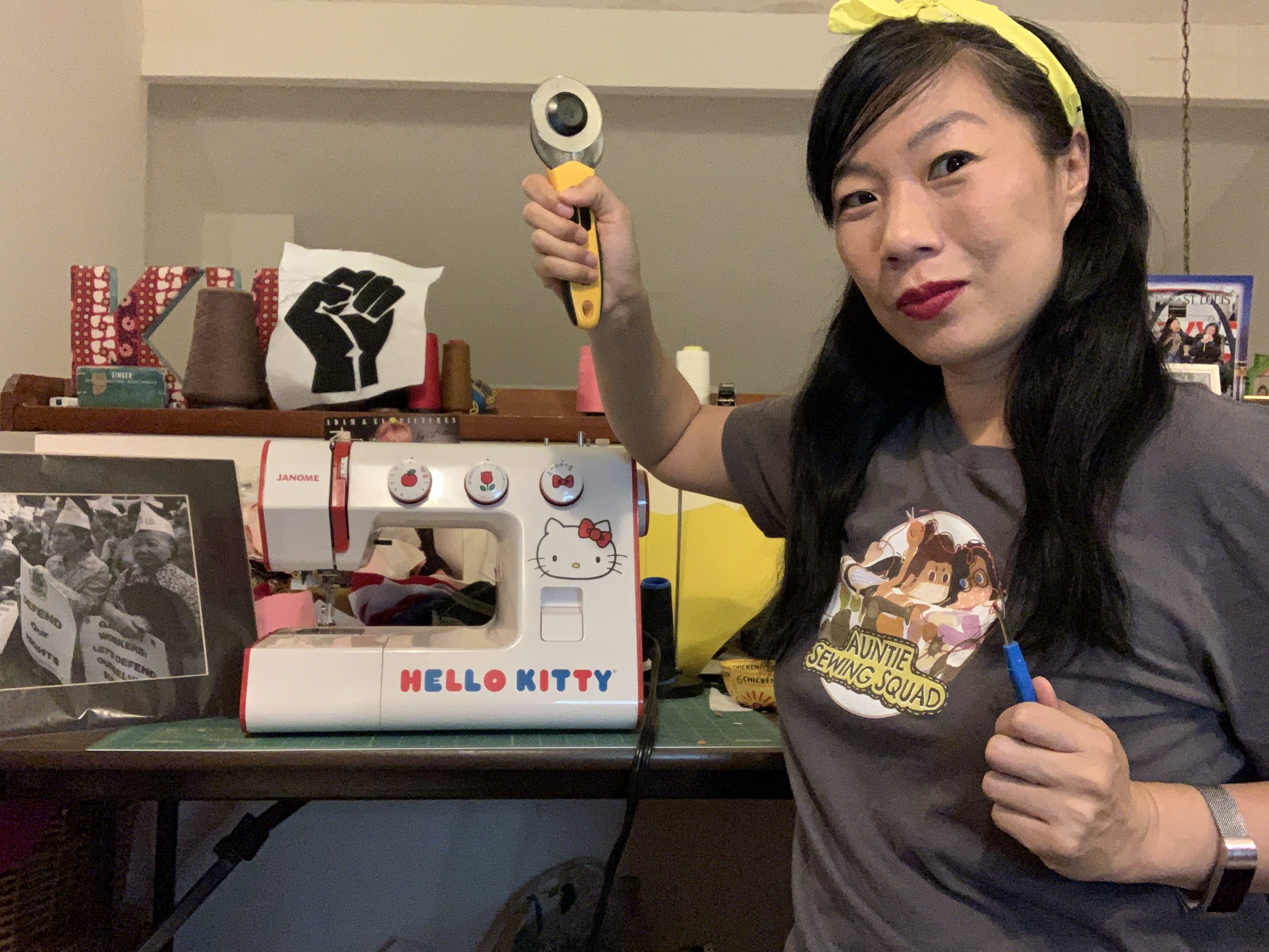 Kristina Wong headshot. In this playful photo, Kristina wields a seam ripper and fabric rotary cutter as she stands next to her Hello Kitty sewing machine in her home studio where her journey with mask making and the Auntie Sewing Squad began in March 2020. Kristina is a middle-aged Chinese American woman with long black hair that is tied back with a yellow bandana. She wears dark red lipstick, a silver watch, and a grey t-shirt featuring the Auntie Sewing Squad logo. Next to her diminutive kiddie sewing machine, is a black and white photograph of elderly Asian American labor protestors by the late Corky Lee. An icon of a Black Power fist is prominently displayed amidst her industrial spools of thread and large red and white patterned stuffed “K” and “W” initials.