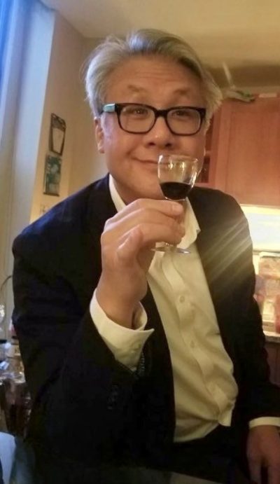 Larry Lee headshot. He is a Chinese American man who self describes his age as a “baby boomer on the verge of retirement.” Larry has short grey hair and is wearing black horn rimmed glasses, a black blazer, a white colored shirt, and he is holding a sherry glass with his pinky extended.