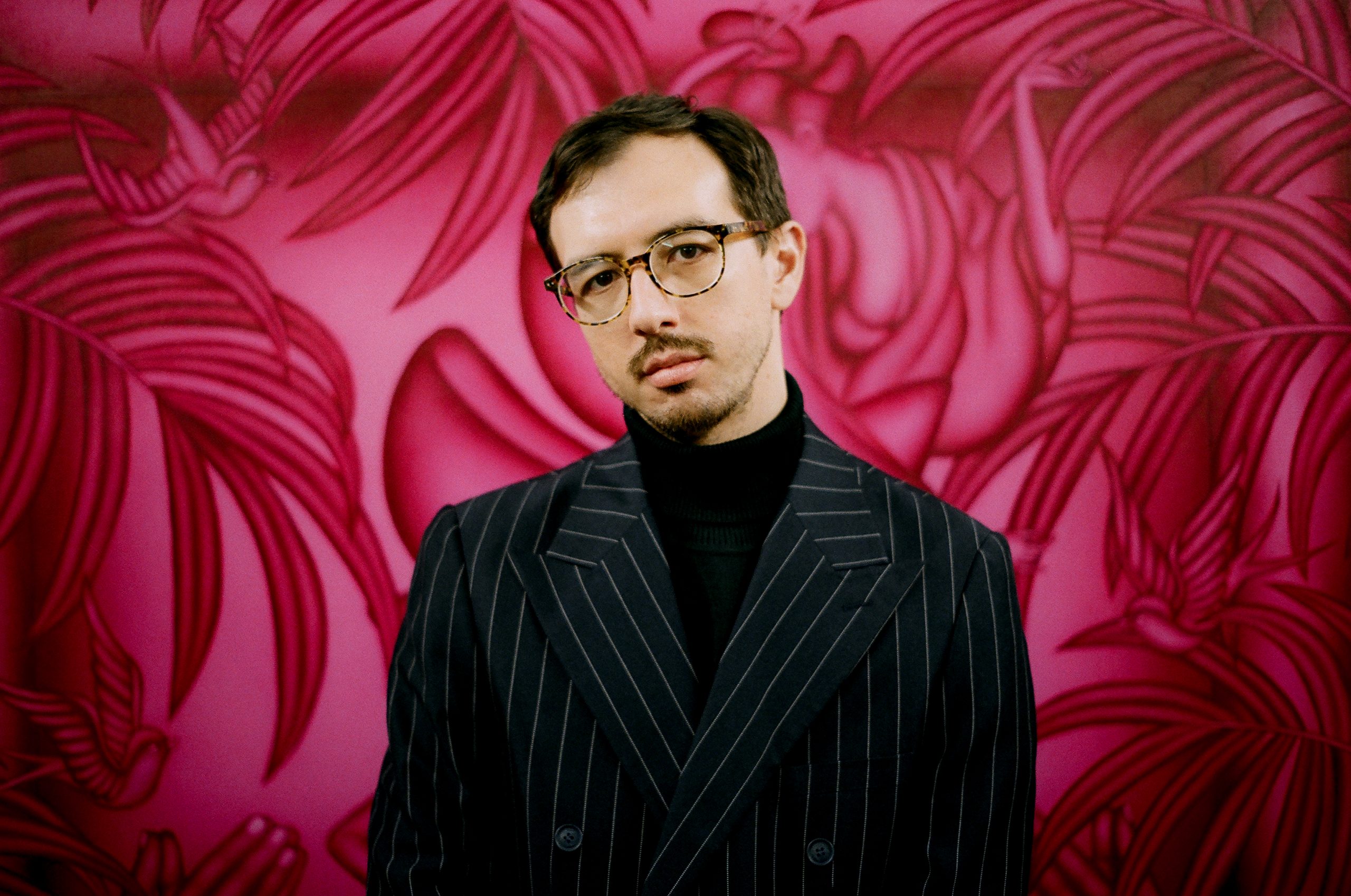 Mathew Tom headshot. In this horizontally oriented photo, a mixed Chinese American man in his mid-thirties stands in front of one of his large-scale magenta paintings. He has his head tilted slightly to the left and looks directly at the camera. Mathew has short brown hair, a goatee, and wears a black turtleneck, a black and white pinstripe blazer with a wide lapel, and tortoiseshell rounded horn-rim glasses. The artist’s painting fills up the entire background with a radiant gradation of fuchsia and black, tropical plant leaves, and birds. Fragments of two figures can be seen as well through folds of fabric, praying hands, and a delicate arm.