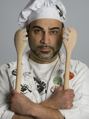 Headshot of Robert Farid Karimi as Mero Cocinero #ThePeoples Cook. A racially ambiguous man with a short black beard and mustache looks directly at the camera with one eyebrow slightly raised. He wears a white chef’s hat, t-shirt with an icon of a raised fist holding a spoon, and a white coat covered in buttons with slogans presumably of leftist resistance. His wrists are crossed against his chest, and he holds wooden salad tongs in each hand.