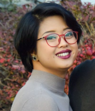Headshot of Mia Matlock. A young non-binary Filipino American person with a black layered bob and carefully manicured eyebrows smiles warmly at the camera in this three-quarter view portrait. She wears a grey mock turtleneck, berry-stained matte lipstick, rounded red horn-rimmed glasses, and round black earrings encircled by diamonds. 