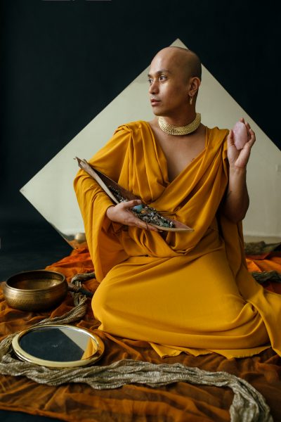 Portrait of Kiam Marcelo Junio. A Filipinx non-binary person with a shaved head is draped in saffron-colored fabric and wears a wide gold choker necklace and gold earrings. They are seated on the floor, with their legs to the side, on top of burnt sienna and orange fabric. Three-quarters of a diamond-shaped mirror is visible in the background. They hold a large pink crystal in their right hand and cradle a coco boat (made from a dried coconut leaf) filled with crystals in their left arm. A silver scarf weaves between an empty hand-hammered brass singing bowl and a gold-rimmed circular mirror on their left side.