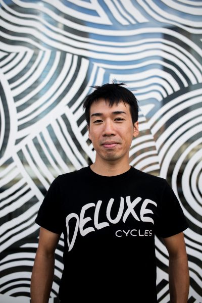 Taro Takizawa headshot. Taro stands in front of one of his black and white patterned murals in this vertically oriented photograph. Taro is a Japanese man in his mid-thirties with short black hair with short layered bangs. He has a slight smile, a 5 O’clock shadow goatee, and he is wearing a black t-shirt with white letterings that reads “Deluxe Cycles.” 