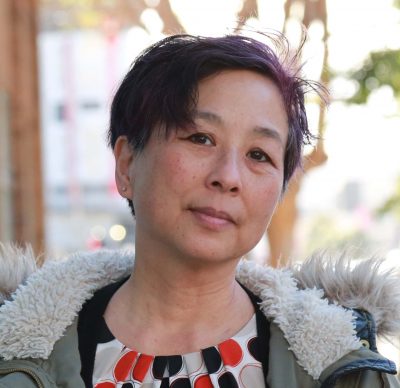Headshot of Valerie Soe against a blurred urban landscape. A Gen X Chinese American woman with her head tilted wears a short purple haircut, polka-dot shirt, and an olive green winter jacket with a fur hood.