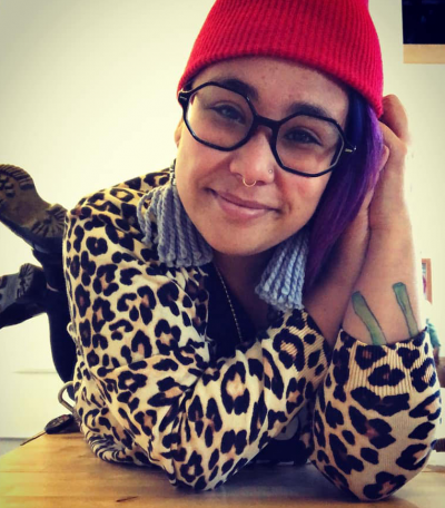 heather c. lou is a racially ambiguous Asian womxn in their 30s. They are laying on their stomach on a wooden table with their legs crossed. They are smiling and leaning their head against their crossed arms. They have long purple hair and are wearing a red knit cap, oversized black hexagon-shaped glasses, a septum ring and nose ring, black boots, black pants, black shirt, and a leopard print cardigan sweater. Part of a tattoo is visible on their left arm.