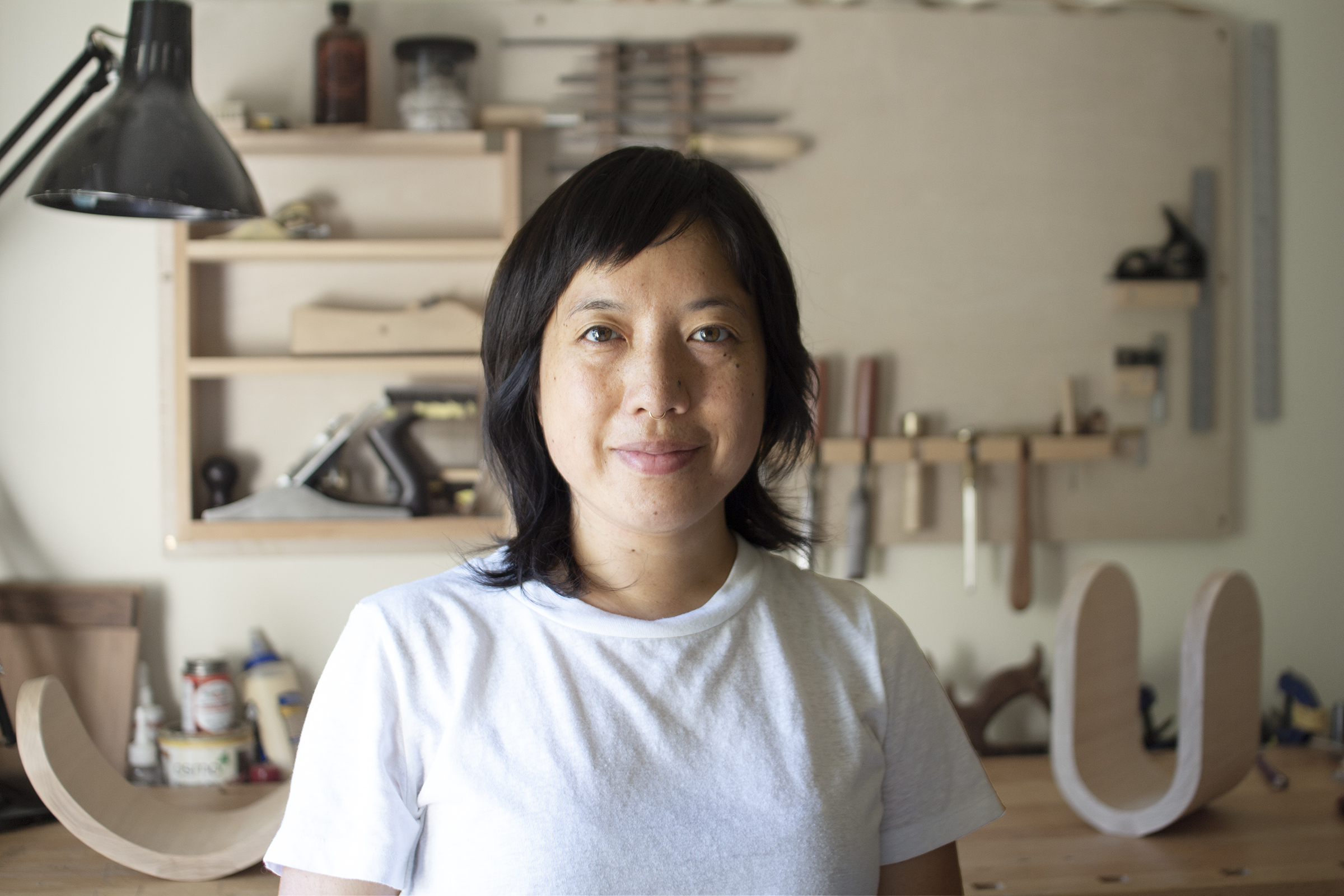 Headshot of Phoebe Kuo. A Taiwanese American woman in her late thirties with shoulder-length fringe cut black hair and a slight smile wears a white t-shirt and gold nose ring as she stands in her sunlit studio. Woodworking tools and works in progress are in the background.